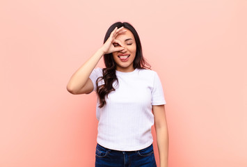 young latin pretty woman smiling happily with funny face, joking and looking through peephole,...