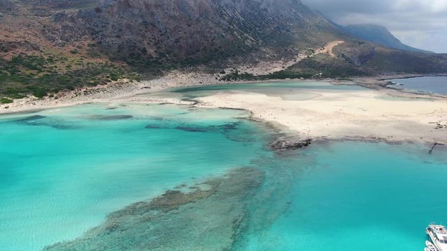 Rocky shore of Balos Beach in Crete Greece with turquoise waters and a tourist transport ship anchored, Aerial flyover reveal shot