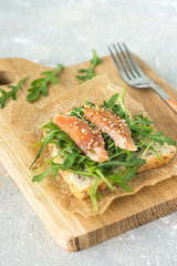 Cereal toast with arugula and trout with sesame seeds on paper and a wooden board top view
