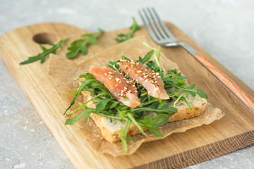 Cereal toast with arugula and trout with sesame seeds on paper and a wooden board