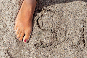 Young girl erases the word summer on the sand. Female foot in the sand closeup