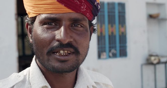Closeup of a happy cheerful positive young brown man with mustache as he smiles looks straight at camera photo video, adjusts his multicolor vibrant turban, outdoors exterior of his small town house