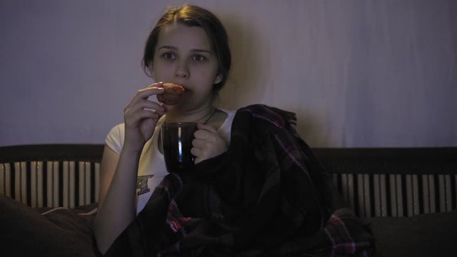 Cute pregnant girl watching tv in a dark room. A pregnant woman in a white T-shirt with the inscription on the chest "baby on board" is watching TV. The girl watches TV and eats.