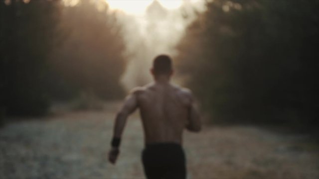 Running sport man. Fit muscular young male runner with naked torso sprinting beetwen trees against dawn, sunrise. Slow motion
