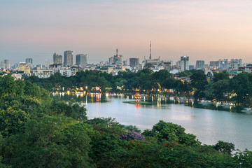 Hoan Kiem lake or Sword lake, Ho Guom in Hanoi, Vietnam with Turtle Tower, green trees and buildings on horizon, at twilight period