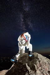 Back view of space traveler reaching top of rocky mountain under mesmerizing night sky with stars, Milky way. Male cosmonaut in space suit exploring new planet. Concept of space travel, cosmonautics