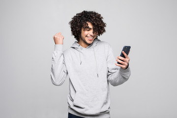 Young curly man playing with the phone looks happy and fisted after winning, isolated on a white background.