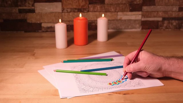 Man painting mandala with color pencils and lit candles over a wooden table. Relaxing paint.