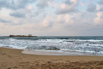 Crete, Greece - A beach with brown sand on the shores of the Mediterranean Sea, waves on the sea, a small church on a small island, clouds in the sky, in the fall in the afternoon.