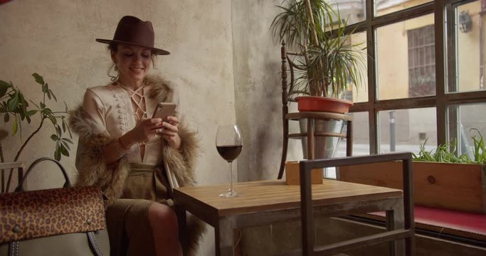 confident strong independent lady girl woman in retro sits in a restaurant with a glass of wine while waiting communicates on the phone happy smiling