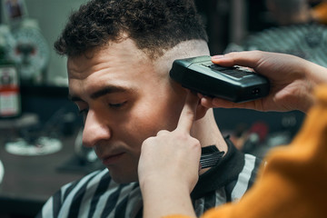 A hairdresser cuts the temple of a curly young man with a hair trimmer.Professional the Barber.the...