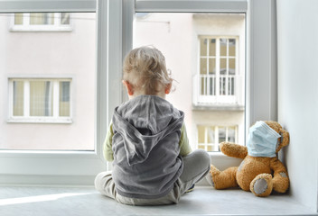 Child in home quarantine looking out of the window with his sick teddy bear wearing a medical mask...