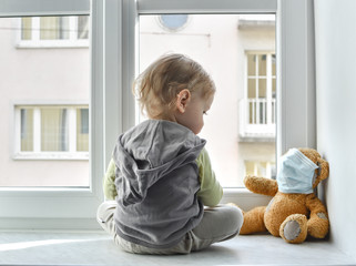 Child in home quarantine standing at the window with his sick teddy bear wearing a medical mask against viruses during coronavirus and flu outbreak. Children and illness COVID-2019 disease concept - obrazy, fototapety, plakaty