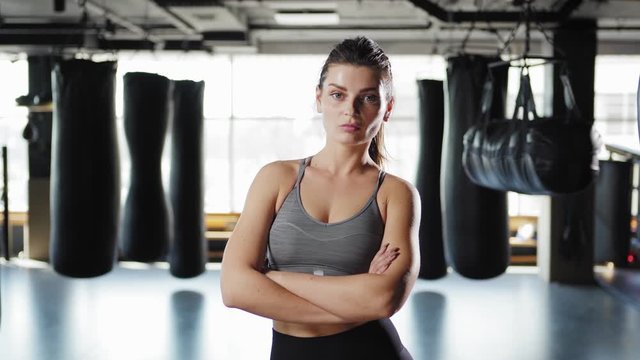 Tilt down waist up shot of fit and confident young woman looking at camera posing cross-armed in boxing gym with heavy bags in background