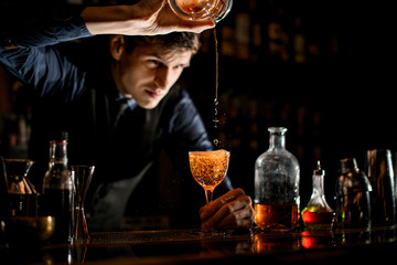 Barman holds wineglass in his hand while pouring a cocktail into it.