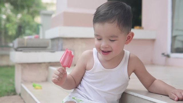 Slow motion video of an Asian toddler boy wearing summer clothes sitting on the stairs outside a house holding a red strawberry popsicle ice cream smiling and thinking
