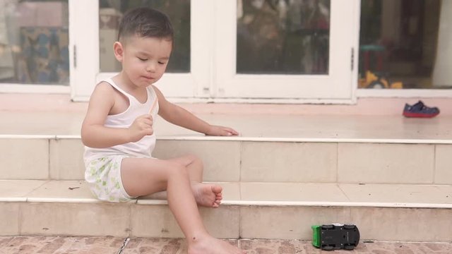 Slow motion video of a handsome Asian boy sitting on the stairs outside the house eating and finishing a popsicle ice cream and spitting it out on the floor.
