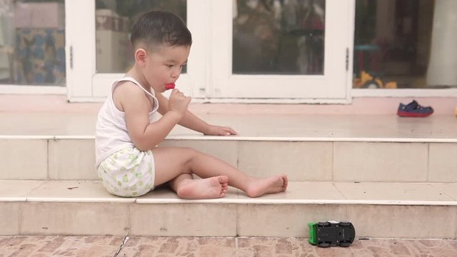 Video of a handsome Asian kid from the side eating a posicle ice cream sitting on the stairs of a house in summer