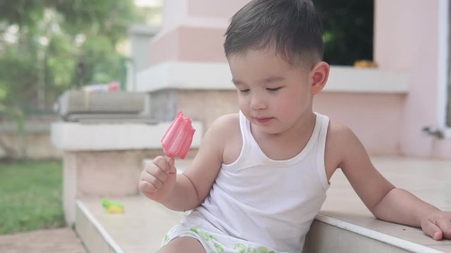 Video of an Asian toddler boy wearing summer clothes sitting on the stairs outside a house holding a red strawberry popsicle ice cream smiling and thinking