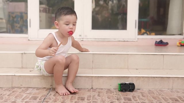 Video of a handsome Asian boy sitting on the stairs outside the house eating and finishing a popsicle ice cream and spitting it out on the floor.