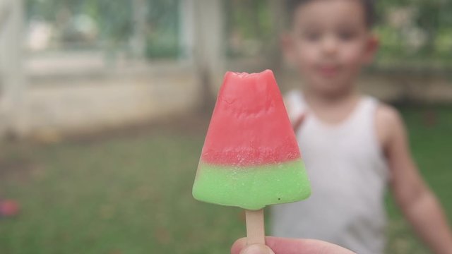 Slow motion video of a parent hand holding a refreshing watermelon popsicle ice cream and Asian boy running to take it and eat it to enjoy a hot summer day