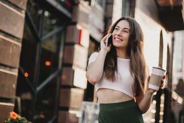 Smiling woman using phone and drink coffee on the street in summer day.