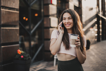 Smiling woman using phone and drink coffee on the street in summer day.