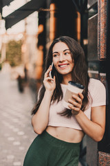 Smiling woman using phone and drink coffee on the street in summer day.