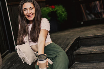 Happy smiling woman drink coffee sitting on wooden steps in street.