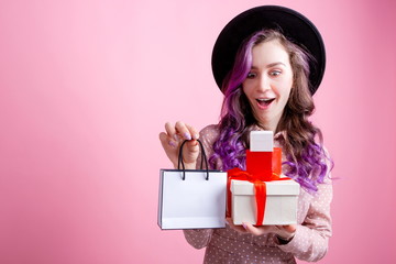 Bright beautiful girl in a polka dot dress standing on a pink background holding gift boxes with a gift bag