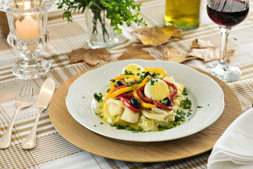 Table set for a dinner, with white decorated portuguese plate. A codfish with bell pepper, boiled egg, black olives and parsley is being served, an typical portuguese dish.