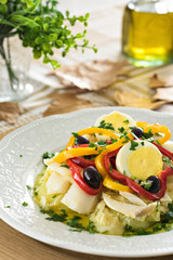 Table set for a dinner, with white decorated portuguese plate. A codfish with bell pepper, boiled egg, black olives and parsley is being served, an typical portuguese dish.