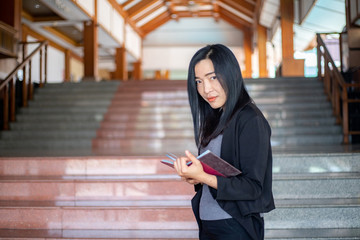 Fototapeta premium The beautiful Asian woman wearing black suit holding book is posing in front of stairs during her working day. 