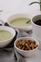 Spring detox broccoli green cream soup in white plates, bowl with crackers and stylish black cutlery on grey background. 