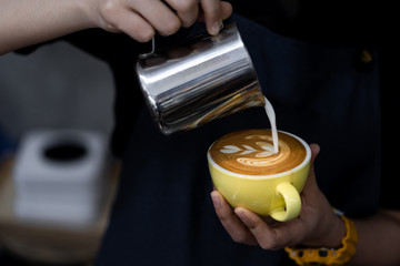 Close-up of barista hand holding and pouring hot milk for prepare latte art on a yellow cup of cappuccino coffee.