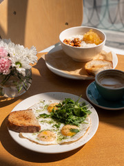 Tasty healthy breakfast: fried eggs, salmon, arugula and croutons on a white plate and  a mug of cappuccino on a wooden table.