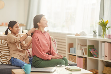 Horizontal shot of happy teen girl sittting on sofa in cozy living room plaiting her mother's hair, copy space