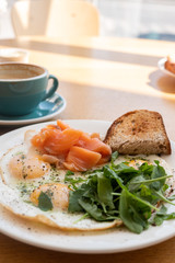 Tasty healthy breakfast: fried eggs, salmon, arugula and croutons on a white plate and  a mug of cappuccino on a wooden table.