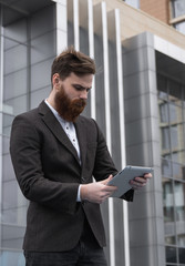 Modern Young bearded Business man working with a digital tablet. Young hipster businessman holding tablet in hands outdoor. Working online with a tablet while standing outside on an office building.