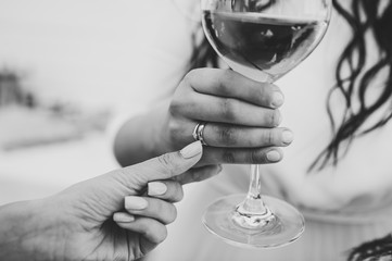 A Black and white photo of lady holding a glass of wine with a wedding ring on finger on party. Girl, woman standing in the background and drink wine. Concept of celebrate, holiday.