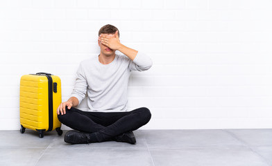 Young handsome man sitting on the floor with a suitcase covering eyes by hands