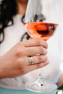 A photo of lady holding a glass of wine with a wedding ring on finger on party. Girl, woman standing in the background and drink wine. Concept of celebrate, holiday.