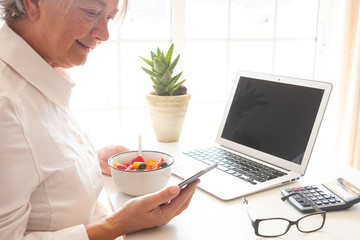 Close up view of a senior woman eating a fruit salad at desk. Computer  on the table - cellphone in the hand - new technology for old people