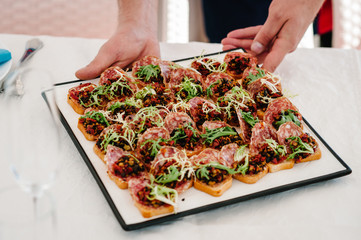 The waiter puts a plate on the table with gourmet appetizers, variety of mini sandwich, crostini with different toppings. Front view above. Assorted Italian appetizer bruschetta.