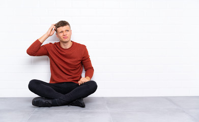 Young handsome man sitting on the floor having doubts and with confuse face expression