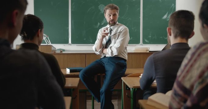 Handsome middle aged male lecturer in shirt sitting on table, holding eyeglasses and talking to students