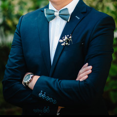 The attractive groom in a suit with bow tie and boutonniere or buttonhole on jacket, is stands on the background greenery in the garden, park. Nature. Cropped photo.