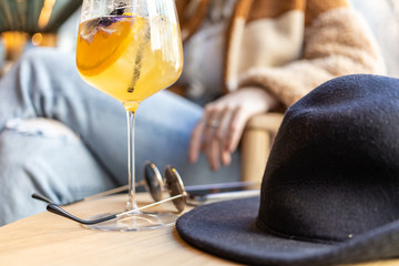 close up view of a woman's hand picking a wine glass filled with delicious natural herbal tea liquor with sprinkles. blurred view of the city buildings with sunglasses and black hat on coffee table