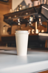 Big white clean cup of coffee to go on the white table. Street coffee, side view. Professional coffee machine at the background blurred Mockup lifestyle concept