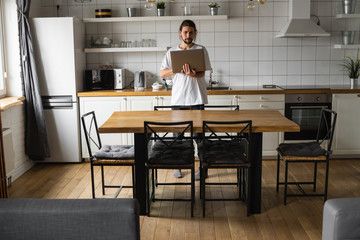 Obraz na płótnie Canvas Freelancer working from home on the kitchen and using laptop. Bearded man working with a laptop and reading good news. Handsome successful self entrepreneur standing with laptop and working at his